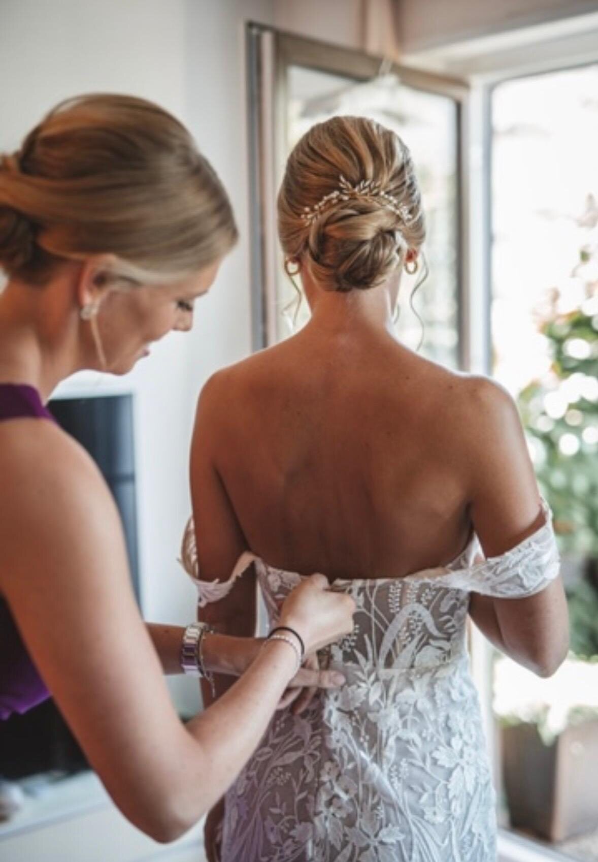 Braut wird beim Getting Ready in ihr Spitzenkleid geknöpft, geflochtene Hochsteckfrisur